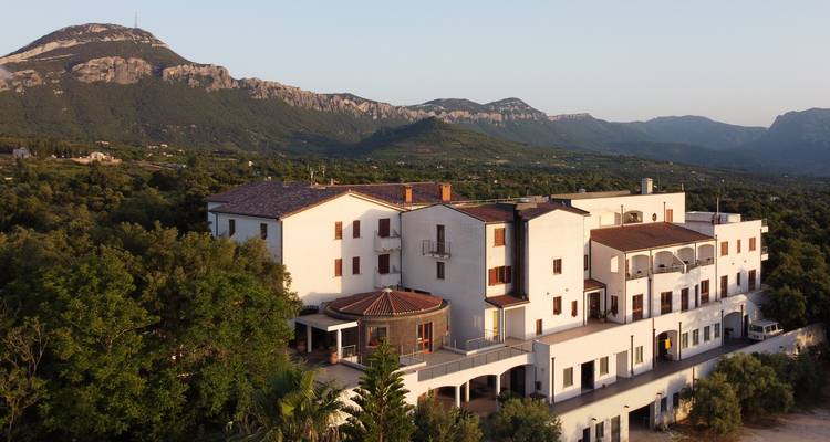 Aerial view of a white hillside hotel complex set against Sardinian mountains.