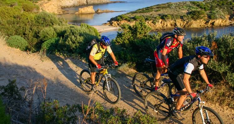 Mountain bikers pedal along a coastal dirt trail overlooking coves and rocky shore.