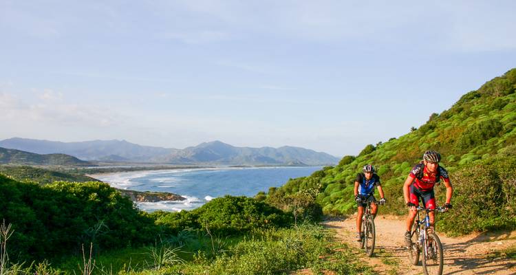 Cyclists ride a rugged coastal path with expansive ocean and mountain views.