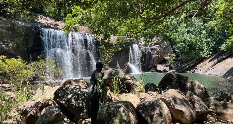 Man standing by a waterfall surrounded by large rocks and trees.