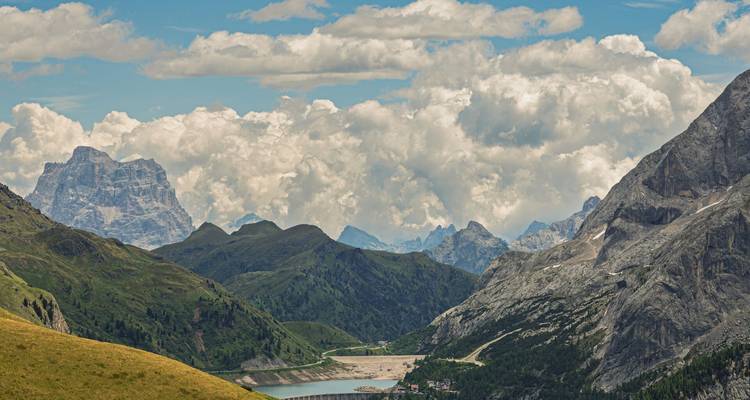 Vue panoramique alpine avec des massifs escarpés, des pentes verdoyantes ondulantes et un réservoir au fond de la vallée.