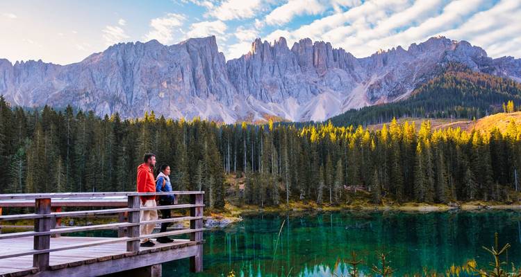 Un couple se tient sur une plateforme en bois surplombant un lac d'émeraude cristallin entouré d'une forêt dense et de pics dramatiques.