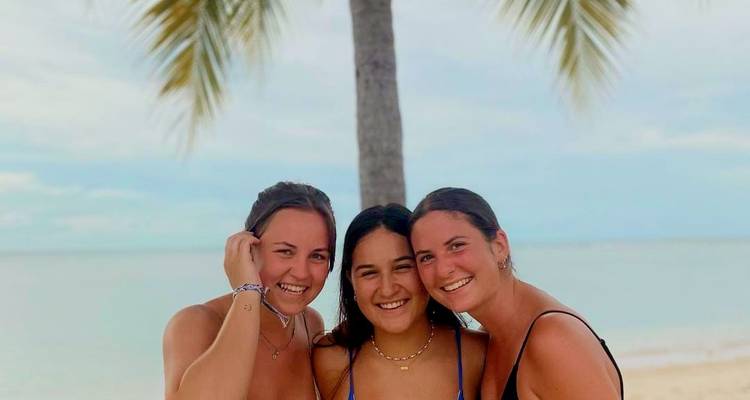 Three women smiling at a beach with a palm tree.