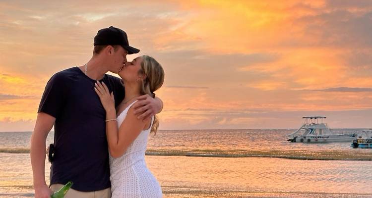 Couple kissing on a beach during sunset with a boat in the background.