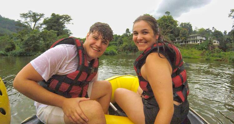 Two people in life jackets smiling in a raft on a river surrounded by tropical vegetation.