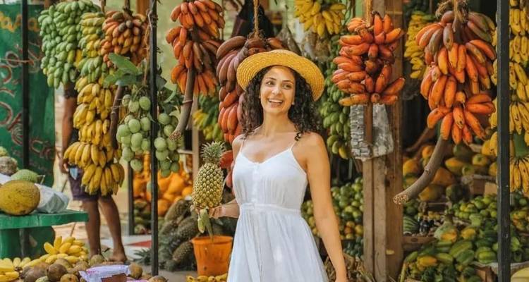 Woman holding a pineapple and standing in front of a fruit stall with bananas.