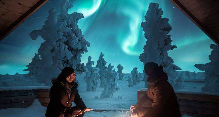 Two people enjoying the Northern Lights from a snowy balcony.