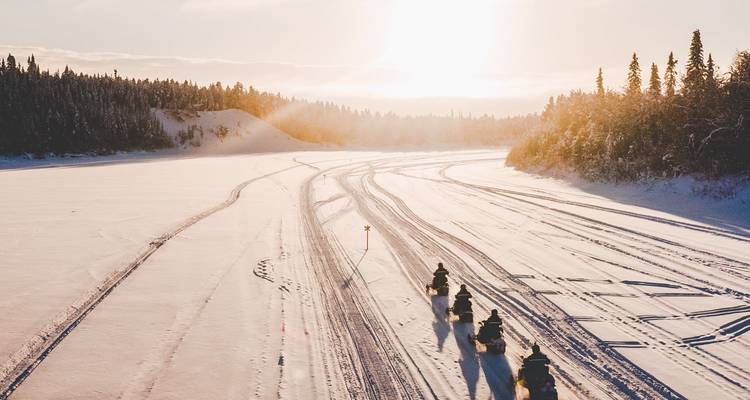 Snowmobile riders driving across a snowy field at sunset.