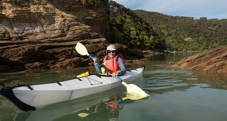 Person kayaking on a calm river with rocky cliffs and forest in the background.