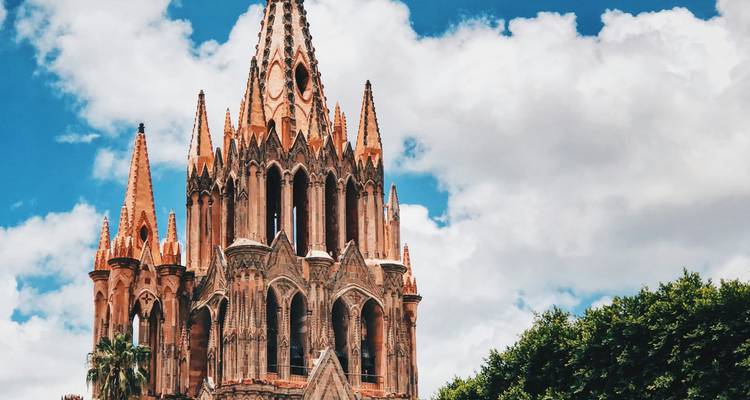 Ornate church spire against a blue sky with some trees visible.