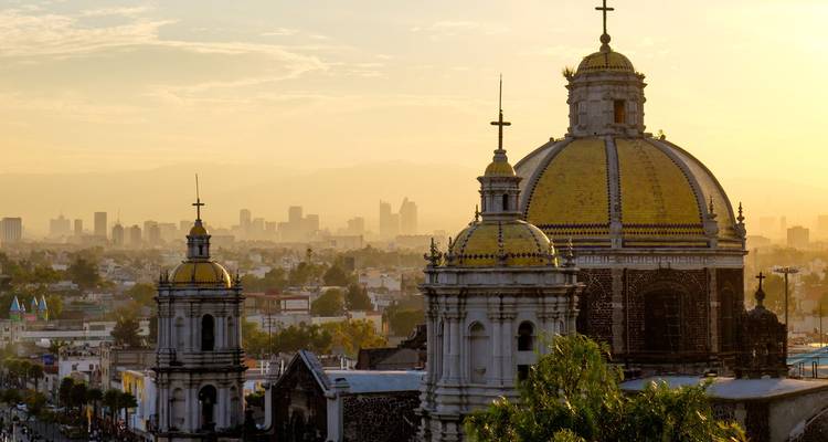 Goudgeel ochtendlicht baadt de koepels van de Basilica de Guadalupe met de skyline van Mexico-Stad op de achtergrond.