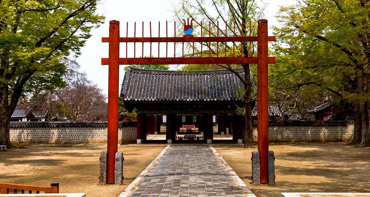 Traditionelles Holztor, das zu einem gefliesten Pavillon führt, der von belaubten Bäumen in Korea umrahmt wird.