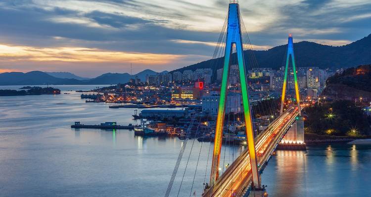 Die beleuchtete Schrägseilbrücke erstreckt sich in der Dämmerung über die Bucht von Yeosu, während Stadtlichter funkeln.