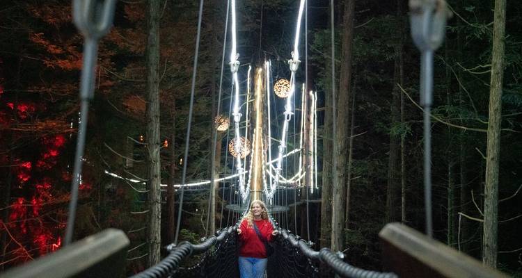 Woman crosses an illuminated forest suspension bridge at night with decorative hanging lights.