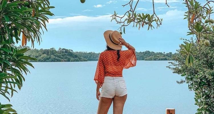 Femme avec un chapeau debout près d'un lac avec des arbres et le ciel en arrière-plan.