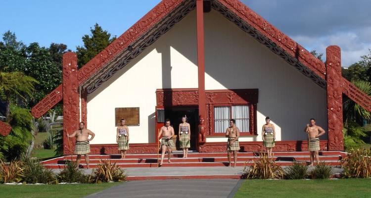 Groupe de personnes en tenue traditionnelle maorie devant une maison de réunion sculptée.