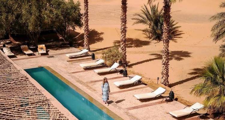 Desert resort pool lined with loungers and palm trees overlooking golden sand dunes, lone woman strolling.