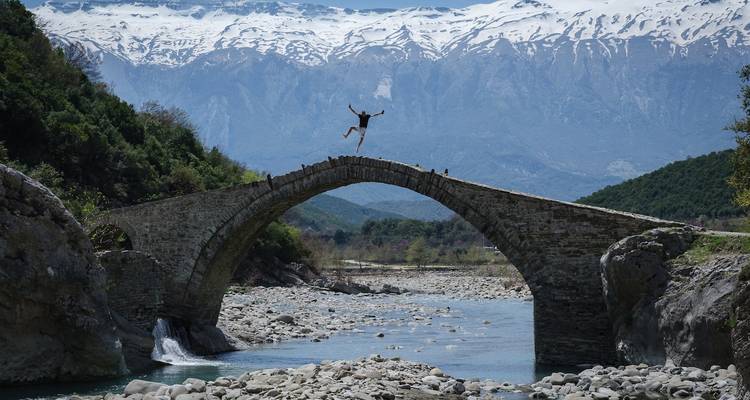 Pont en arc de pierre au-dessus d'une rivière avec des montagnes à l'arrière-plan.