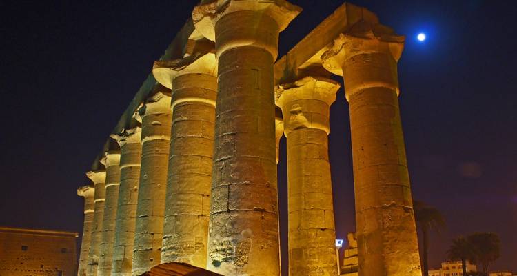Massieve stenen zuilen van de Luxor Tempel verlicht door amberkleurige lichten tegen een nachtelijke hemel met heldere maan.