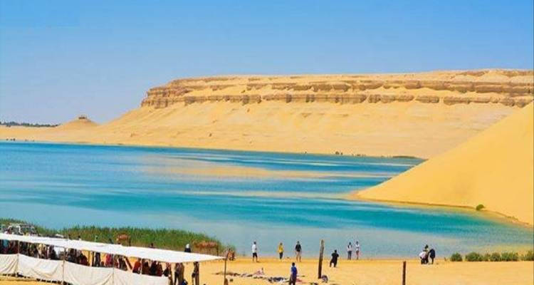 Lago turquesa del desierto bordeado por dunas doradas y acantilados con visitantes paseando bajo sombrillas.