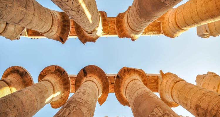 Blik omhoog naar rijen van massieve zandstenen zuilen in de Karnak Tempel tegen een lichtblauwe hemel.