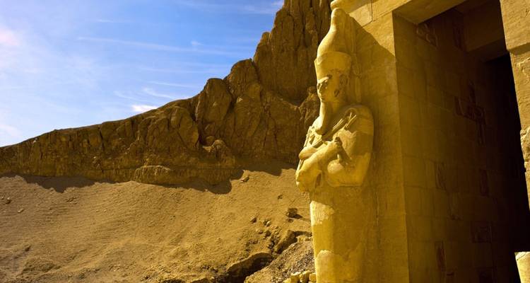 Gemeißelte Pharaonenstatue, die einen steinernen Türeingang neben kahlen Wüstenklippen bewacht.