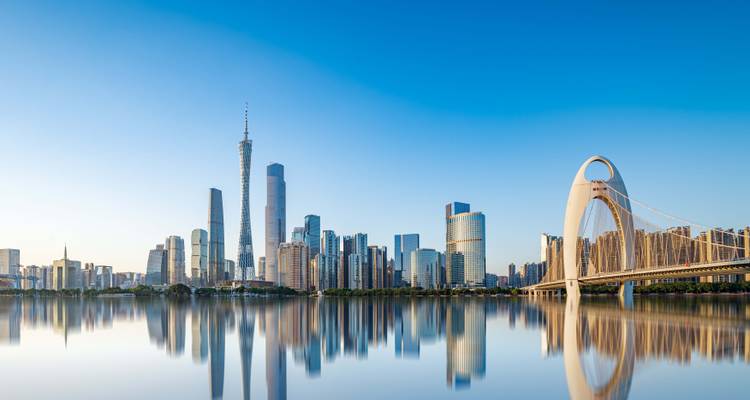 A cityscape of modern skyscrapers with a large bridge reflecting on calm water, under a clear blue sky.