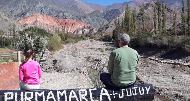 Adult and child sit on a rock wall overlooking the multicolored hills of Purmamarca valley.