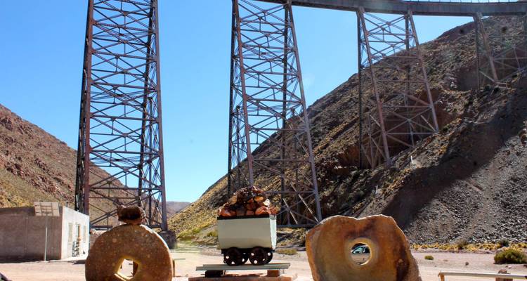 Tall iron railway viaduct towers above a desert canyon with a small mining cart display below.