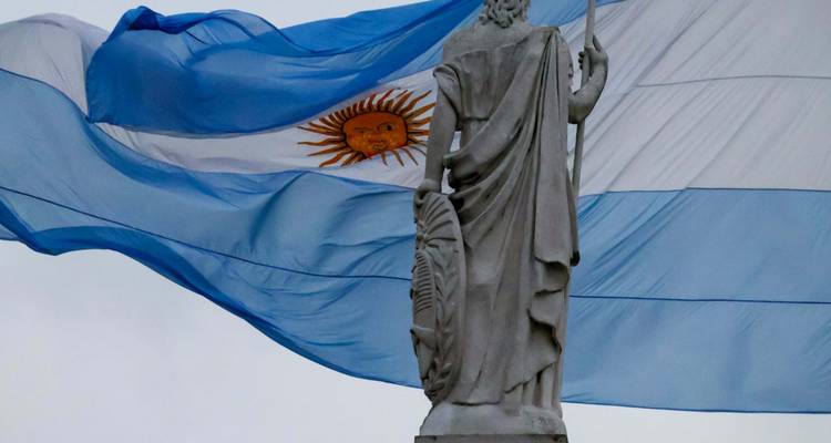 Stone statue seen from behind with a large Argentine flag flowing dramatically in the wind.