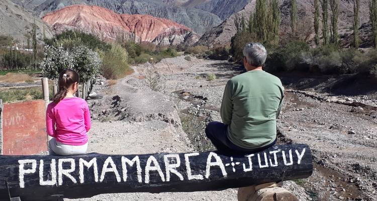 Father and daughter sit on a beam painted Purmamarca-Jujuy facing colourful hills and dry riverbed.