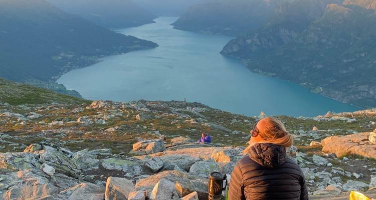 Un senderista con una chaqueta acolchada se sienta en las alturas rocosas mirando hacia un fiordo enorme bañado por la suave luz del atardecer.