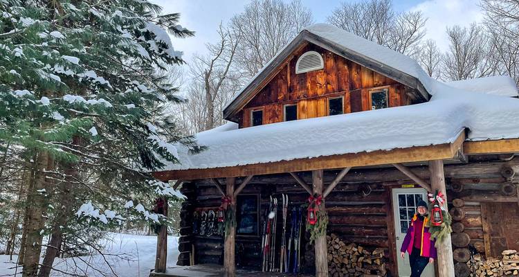 Cabane en rondins dans une forêt enneigée avec une personne debout sur le porche.