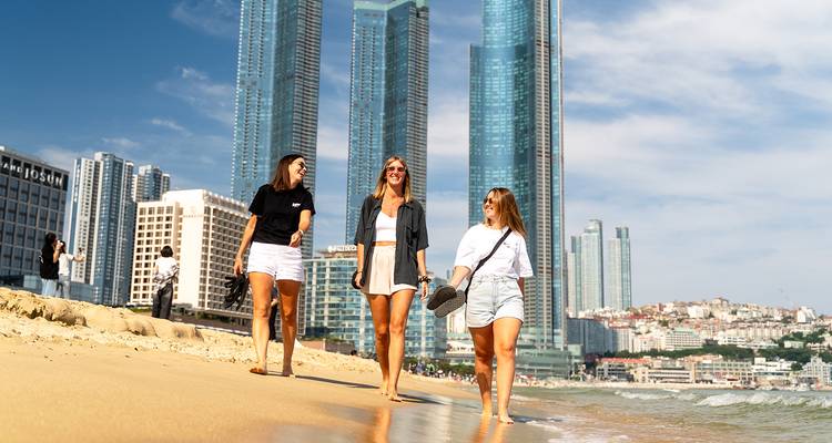 Trois femmes marchant sur une plage avec de grands bâtiments en arrière-plan.