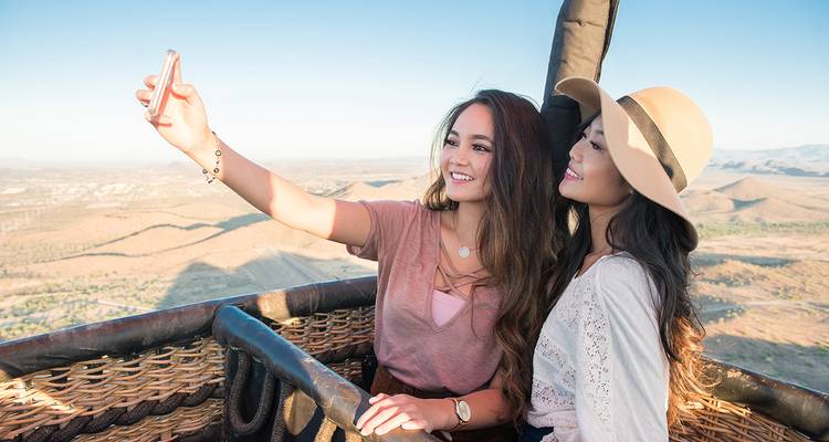 Deux femmes prenant un selfie dans une montgolfière sur fond de paysage.