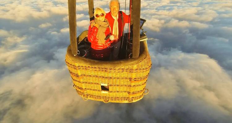 Un couple dans une montgolfière au-dessus des nuages, portant des vestes rouges.