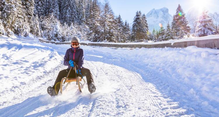 Person sledging down a snowy hill with mountains in the background.