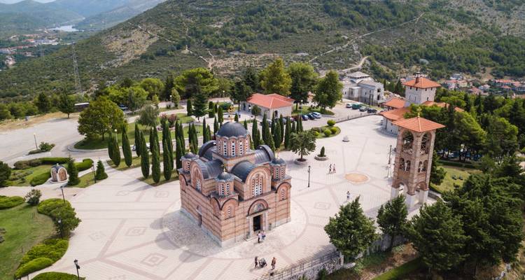 Elevated view of a Serbian Orthodox monastery complex with striped brick church, cypress trees and bell tower set on a hillside.