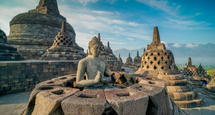 Statue sereine de Bouddha assise parmi les anciens stupas de pierre du temple de Borobudur au lever du soleil avec des montagnes brumeuses au-delà.