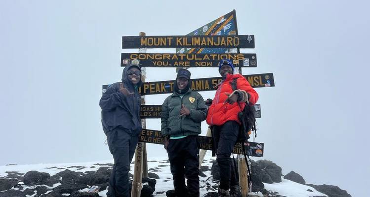 Trois personnes posant au sommet du mont Kilimandjaro à côté d'un panneau.