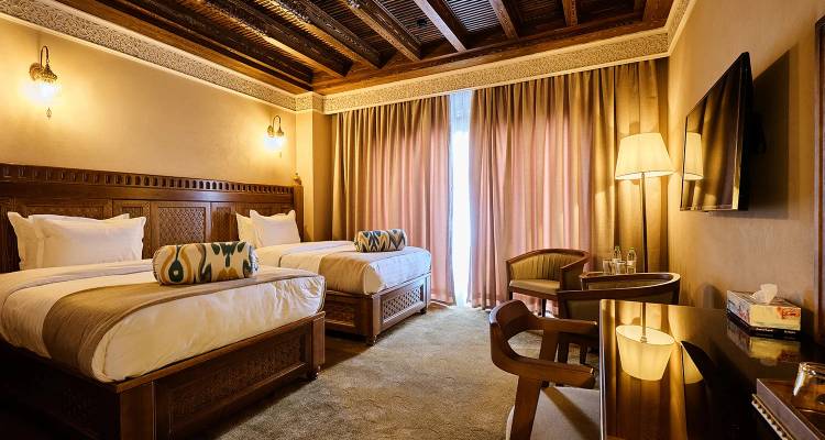 Elegantes Hotelzimmer mit zwei Einzelbetten, geschnitzten Holzkopfteilen und warmer Umgebungsbeleuchtung.