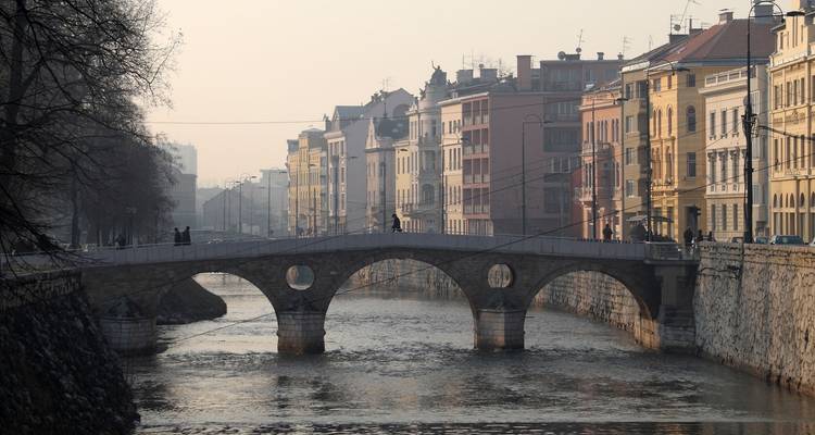 Puente de arco de piedra cruzando el río Miljacka con edificios de colores pastel alineados en el malecón bajo la suave luz de la mañana.