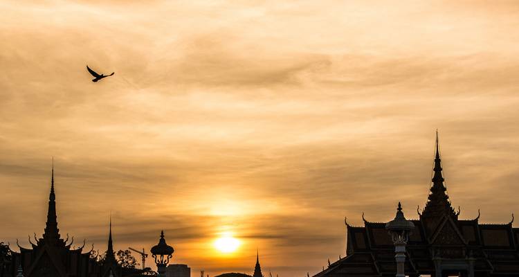 Silhouetted Khmer palace spires and a flying bird against a dramatic orange sunset sky.
