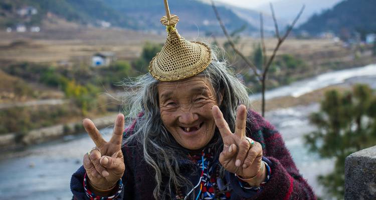 Elderly woman with traditional hat making peace signs by a river.