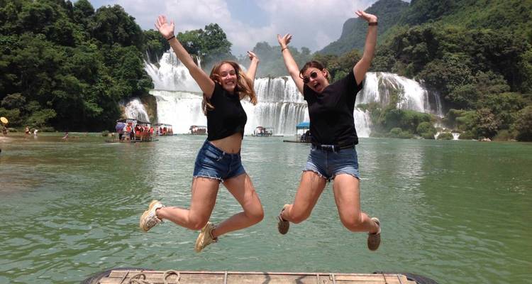 Deux jeunes voyageurs sautent joyeusement devant une grande cascade et une rivière émeraude.