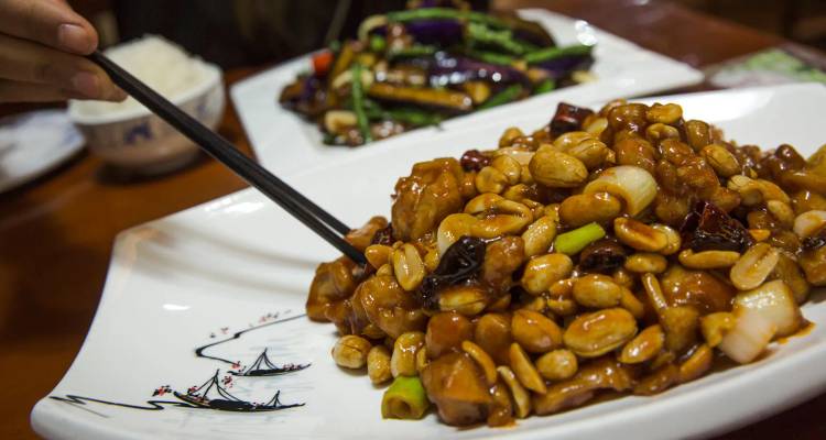 Close-up of a saucy peanut-topped stir-fry being picked up with chopsticks in a restaurant.