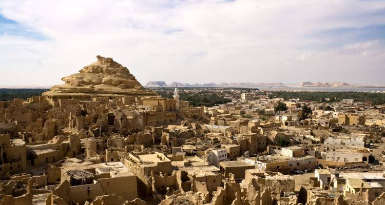 Panoramic view over the mud-brick ruins of Shali Fortress rising from the Siwa Oasis with desert landscape beyond under a bright blue sky.