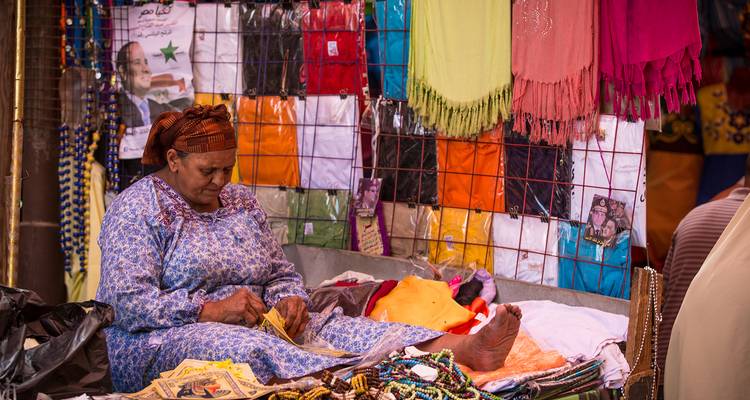Femme assise à un étal de marché avec des foulards colorés et des colliers de perles.