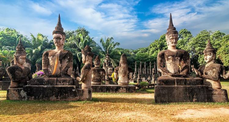 Steinerne Buddhastatuen mit spitzen Stupas, aufgereiht im Buddha-Park unter blauem Himmel und Palmen.