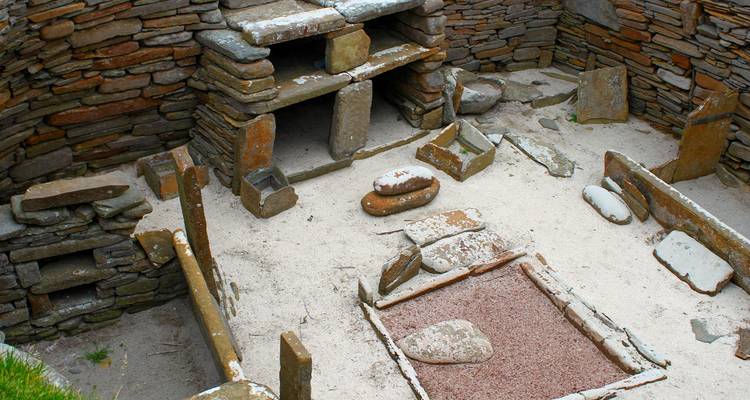 Steinernes Inneres einer neolithischen Behausung in Skara Brae mit erhaltener Feuerstelle und Möbeln.